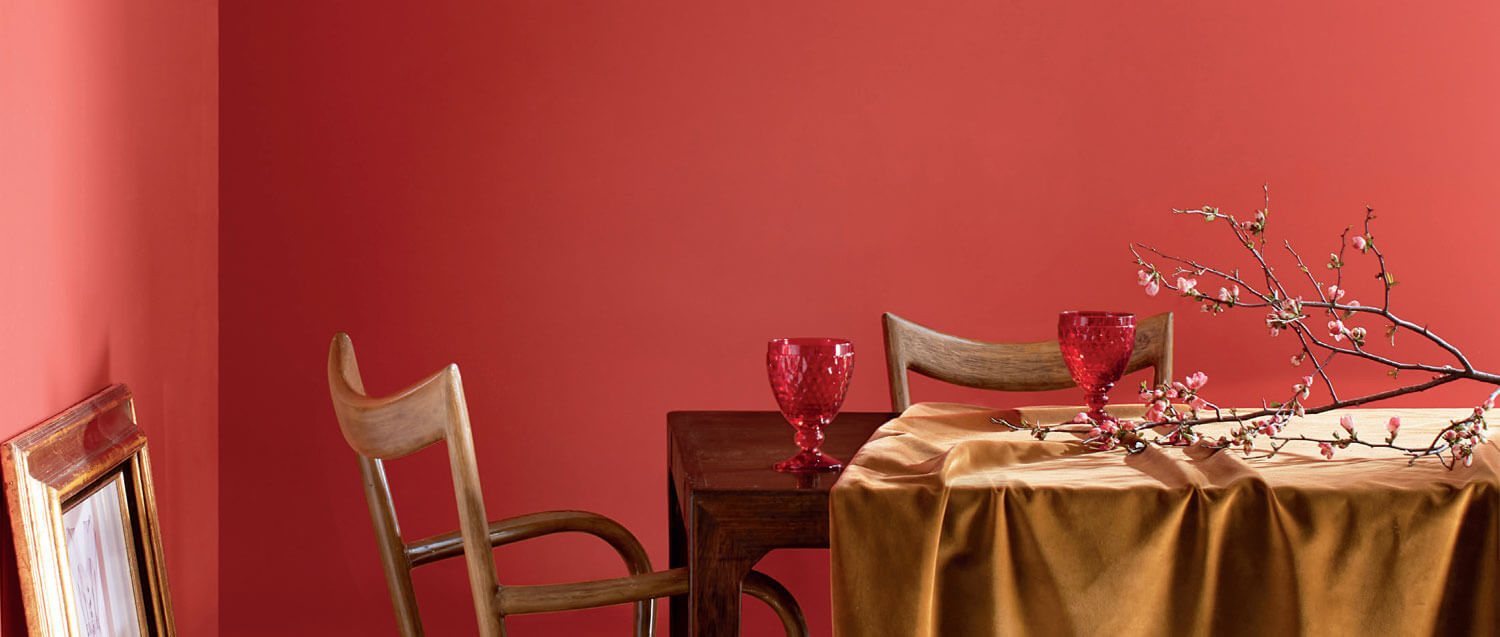 Dining room with coral tinged with pink painted wall, a table, chairs, cups and a branch with buds